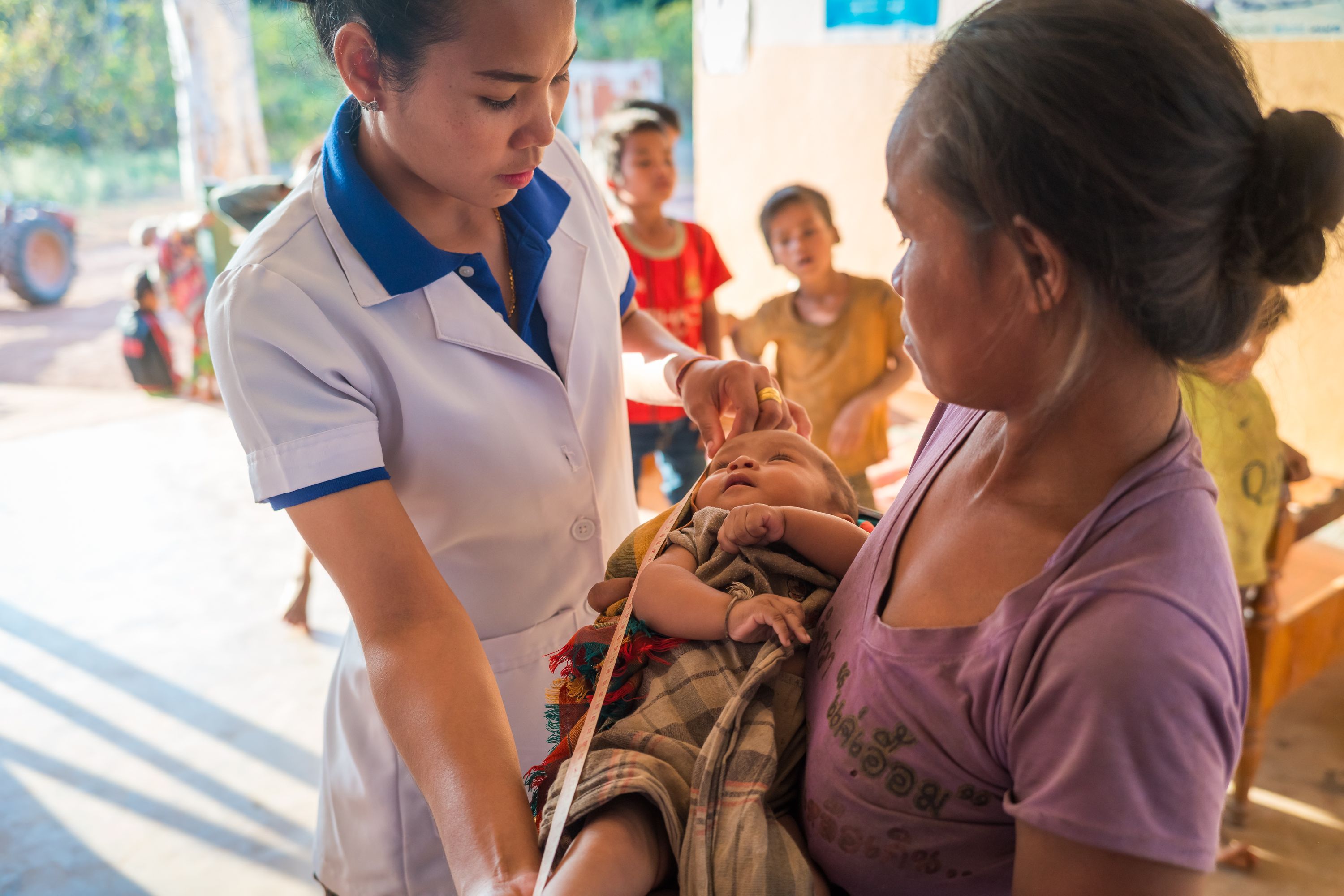 A community health worker measuring a baby while the mother holds the child at a rural health clinic in Southeast Asia
